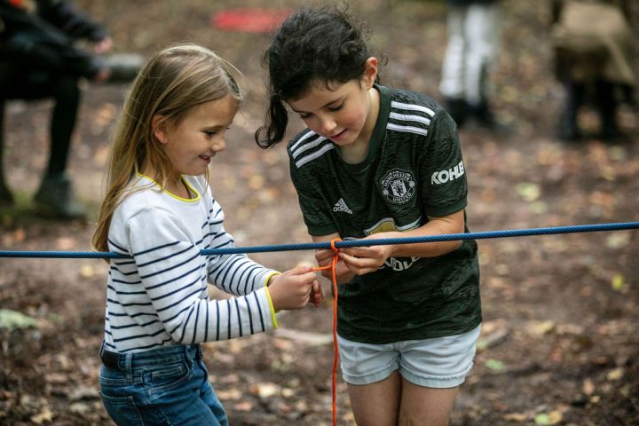 Two children tie a rope in a forest setting, smiling and concentrating on the task.