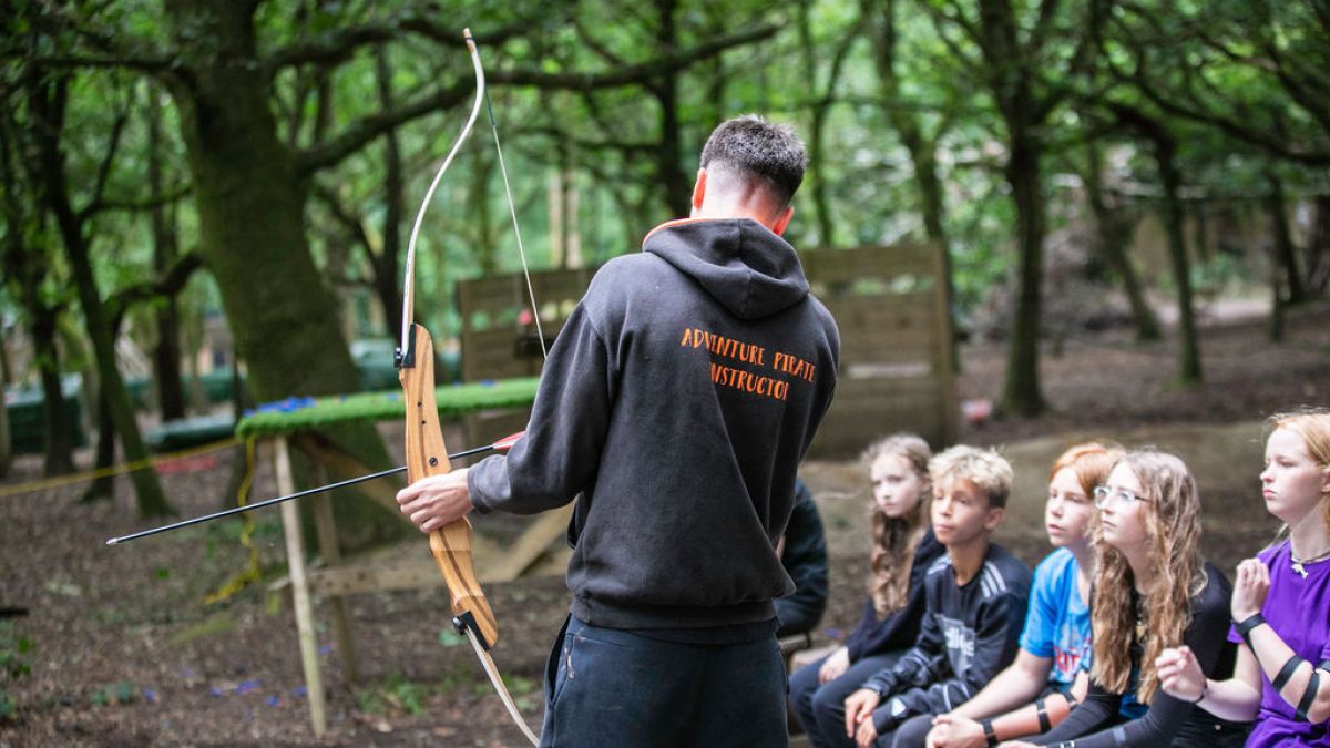 Instructor with bow teaches group of kids in forest setting.