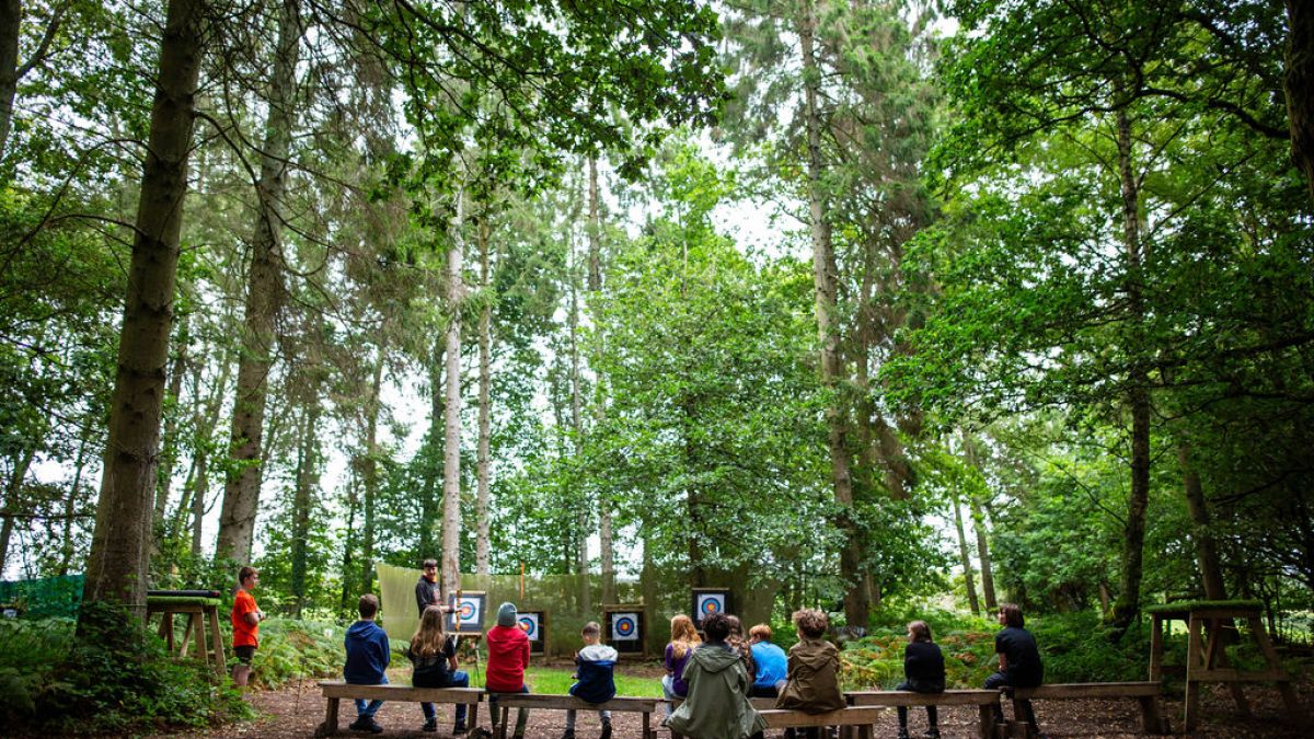 Children seated on benches in a forest observing archery targets.