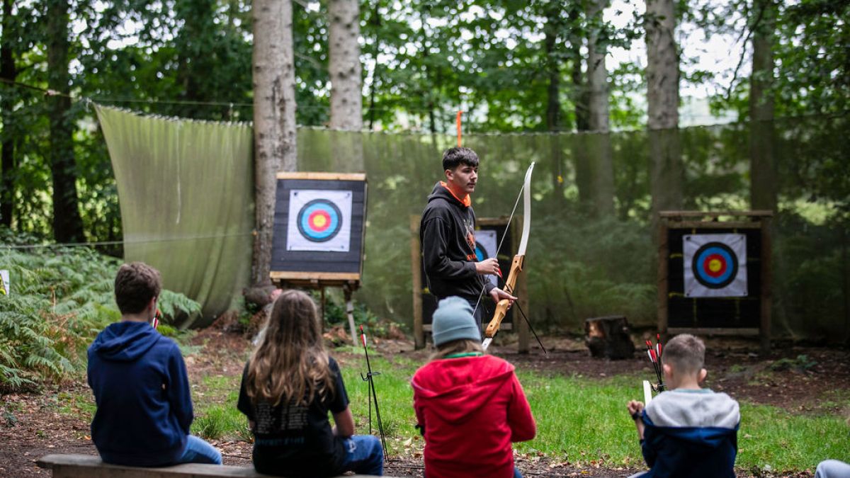 Instructor with bow teaches archery to children seated outdoors with targets nearby.