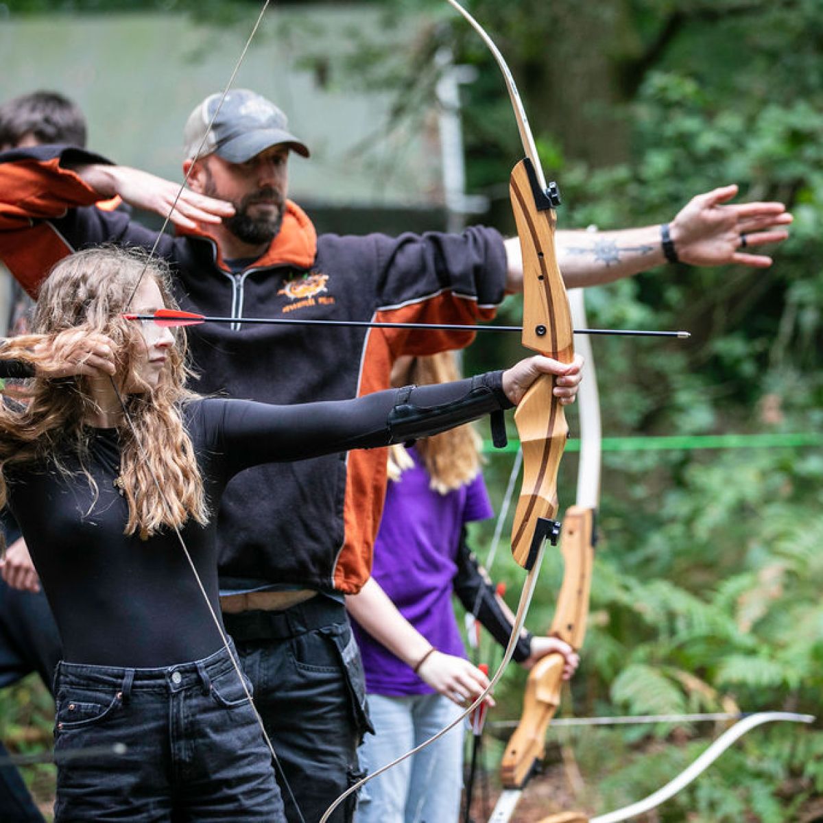 Two people aiming bows and arrows in an outdoor archery setting, surrounded by greenery.