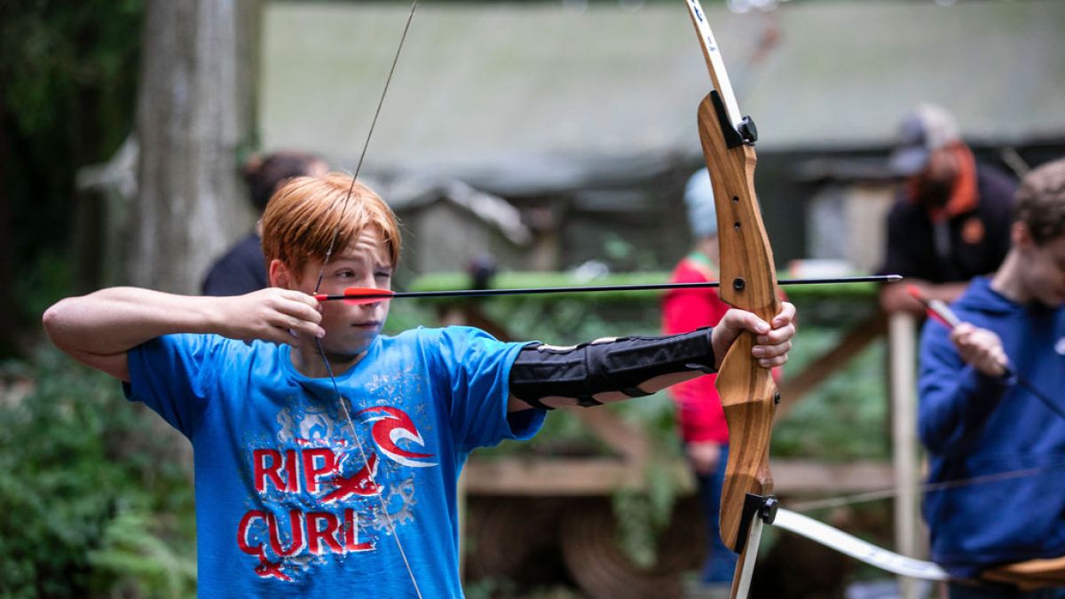 Person aiming a bow and arrow in an outdoor archery setting with others in the background.