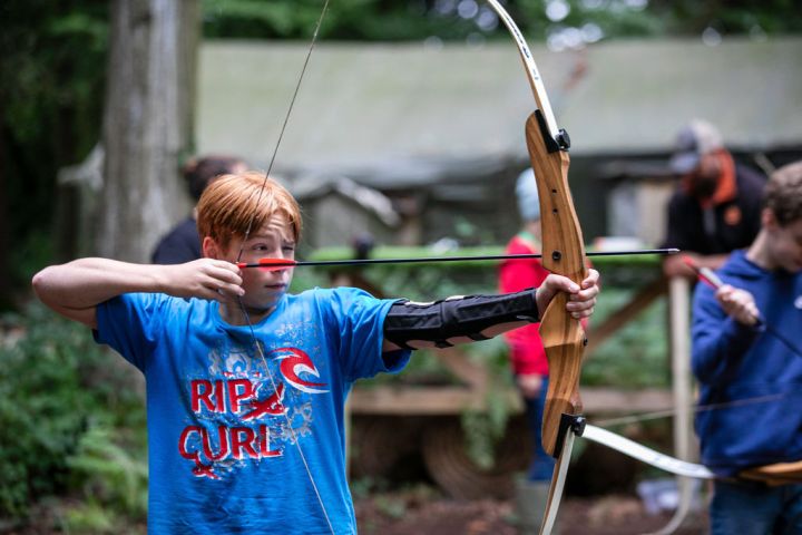 Person aiming a bow and arrow in an outdoor archery setting with others in the background.