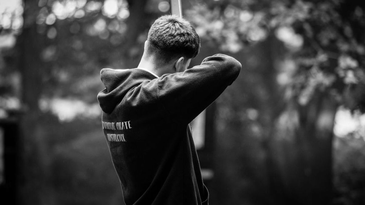 Person in a hoodie holds a stick outdoors in a forest setting, viewed from the back.