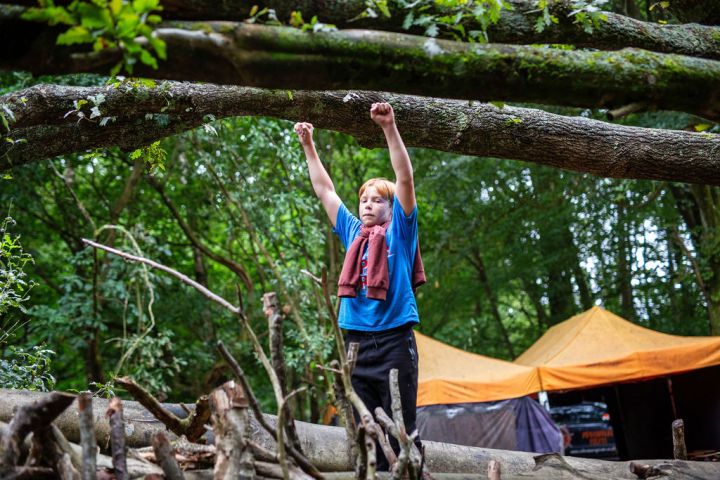 Child in blue shirt climbs tree in forest with orange tents in background.
