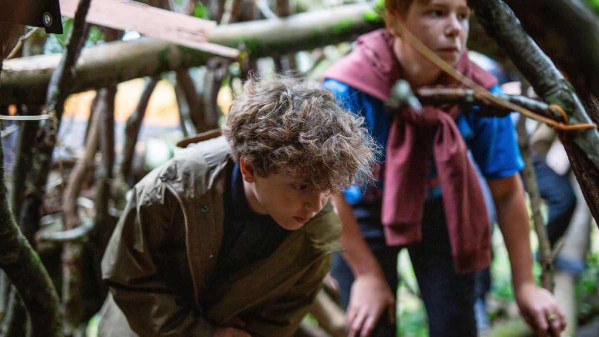Two kids exploring a wooden shelter made of branches in a forest.