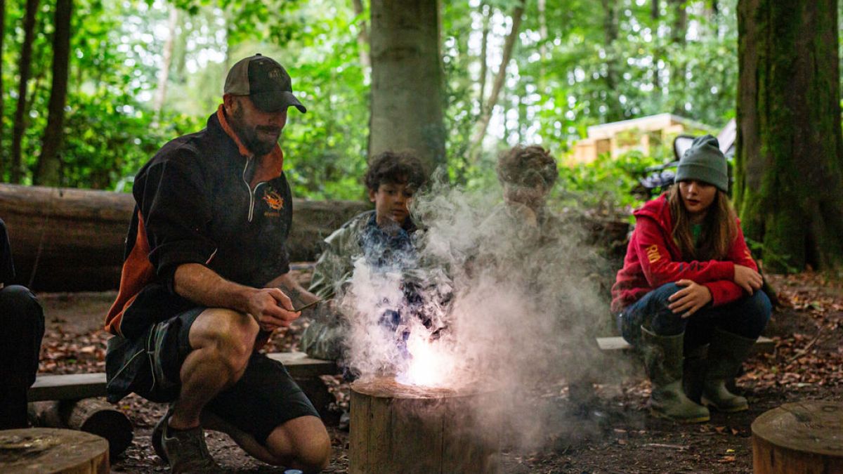 Group of people gathered around a fire in a wooded area with smoke rising.