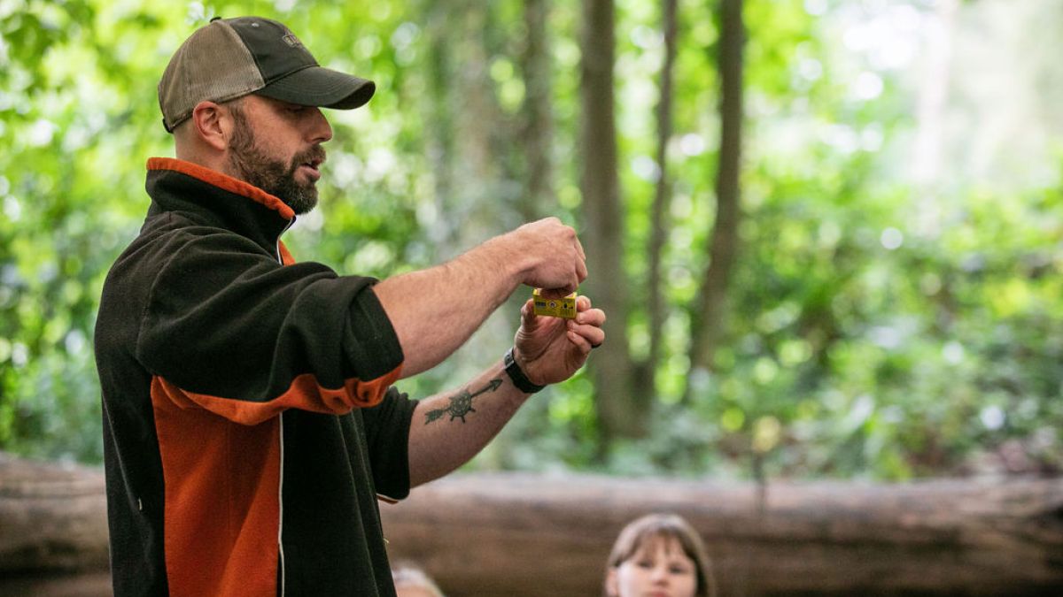Man outdoors demonstrating with small object, children watching.