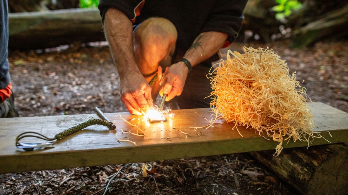 Person using flint to start a fire with kindling on a wooden bench outdoors.