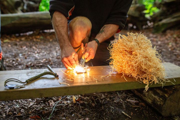 Person using flint to start a fire with kindling on a wooden bench outdoors.