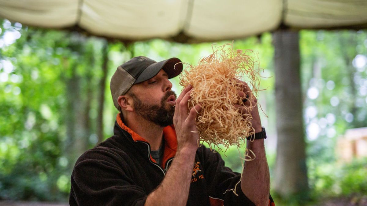 Man outdoors blowing on a bundle of straw in a forest setting.