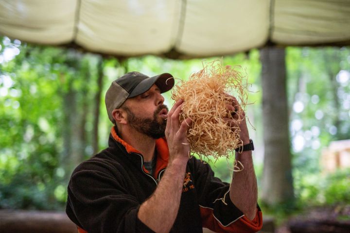 Man outdoors blowing on a bundle of straw in a forest setting.