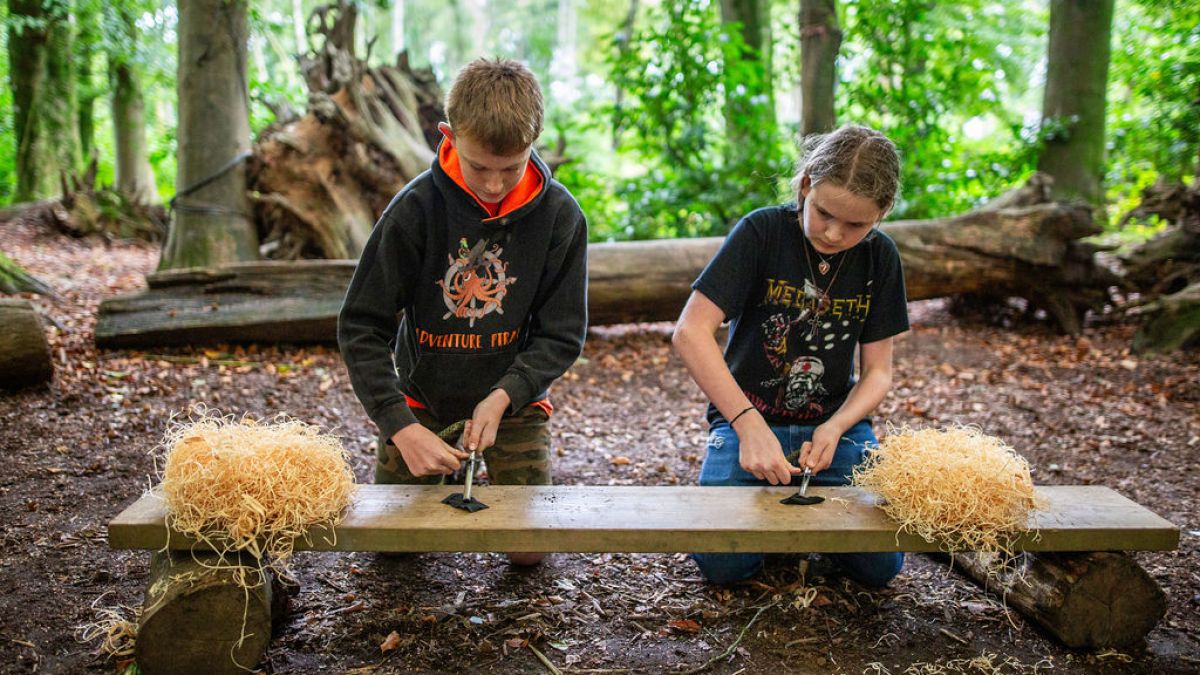 Two kids in a forest practice fire-starting techniques with tools and wood shavings on a wooden bench.