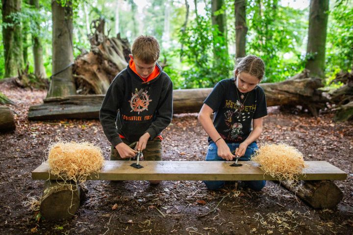 Two kids in a forest practice fire-starting techniques with tools and wood shavings on a wooden bench.