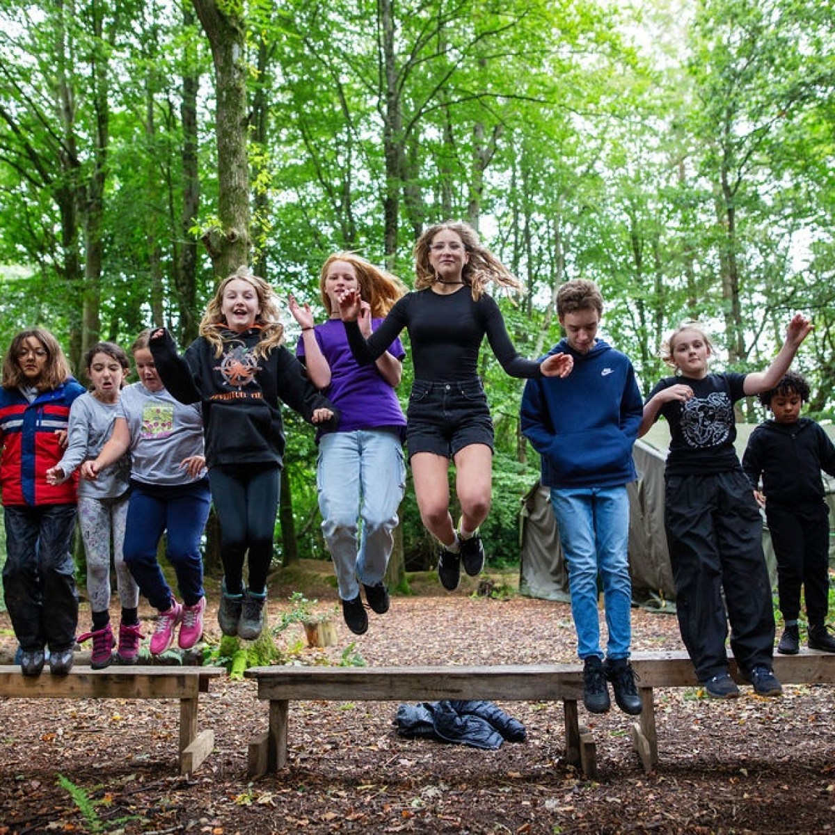 Group of kids jumping off benches in a forest setting.