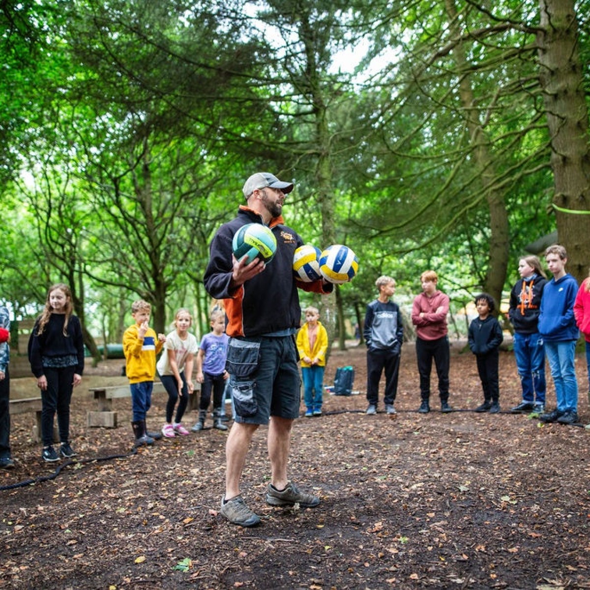 Instructor with volleyballs talking to children in a forest clearing.