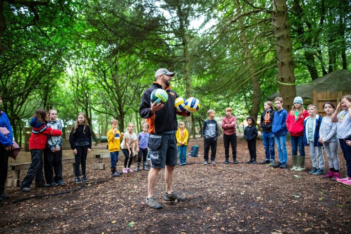 Instructor with volleyballs talking to children in a forest clearing.