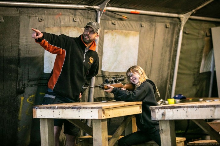 Person instructs a girl aiming a rifle inside a tent with wooden tables.