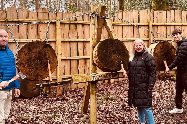 Three people inside a wooden enclosure holding axes near tree trunk targets.