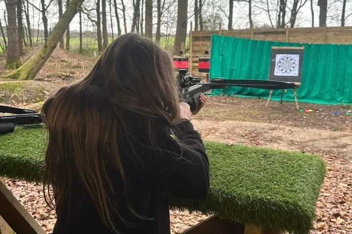 Person with long hair aiming a rifle at a target in an outdoor shooting range.