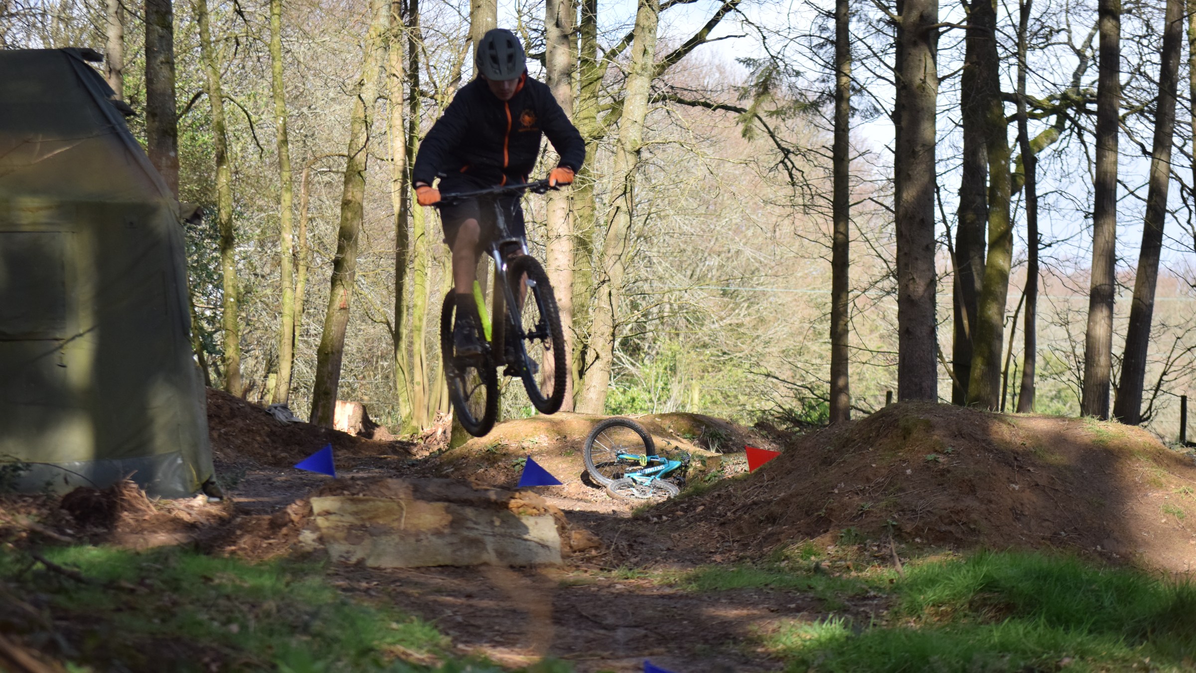 Person mountain biking, mid-air jump in a forest trail setting.
