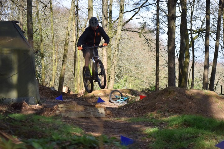 Mountain biker jumping over a dirt ramp in a wooded trail.