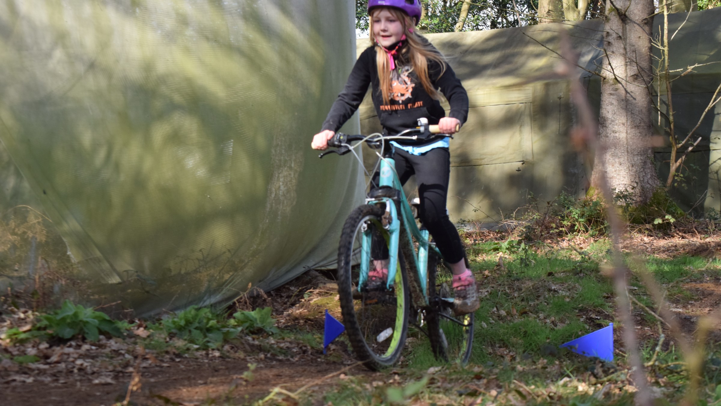 Child in purple helmet riding a bike on a forest trail with green netting in the background.