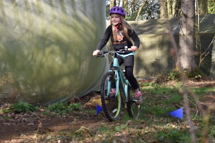 Child in purple helmet riding a bike on a forest trail with green netting in the background.