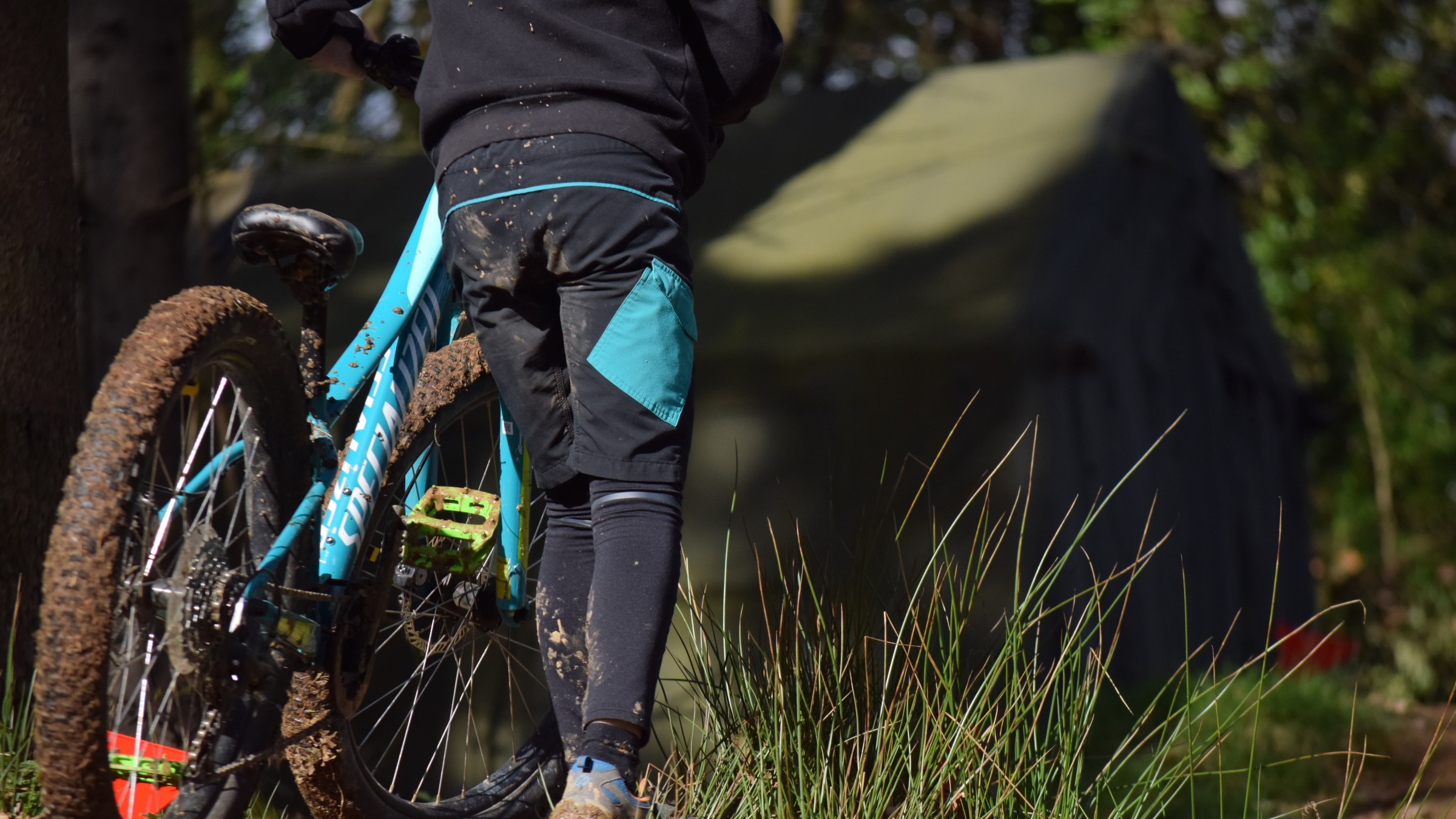 Person with muddy mountain bike stands near a tent in a forest clearing.