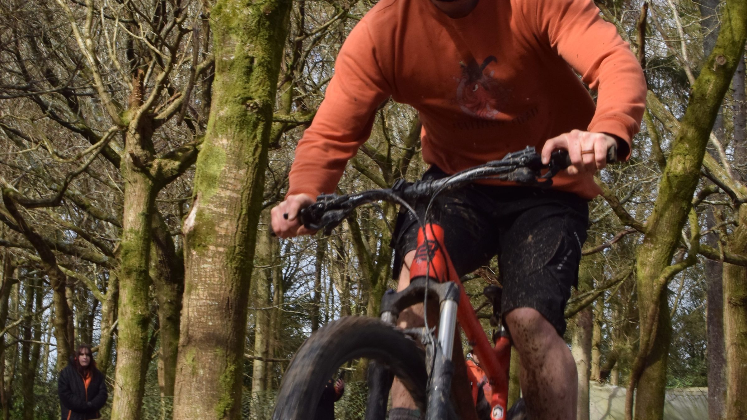 Person in orange helmet mountain biking in a forest, jumping off a small ramp.
