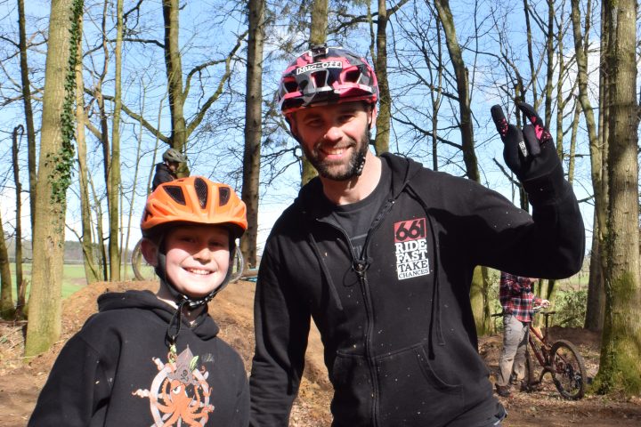 Two people smiling in helmets, posing in a forest with a bicycle in the background.