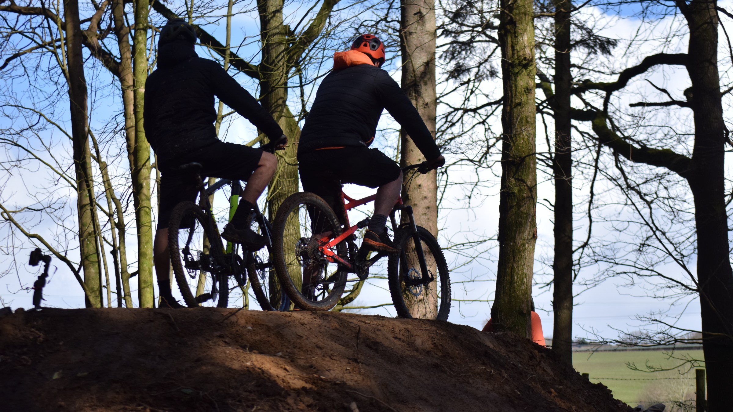Two mountain bikers on a dirt path in a wooded area, viewed from behind.