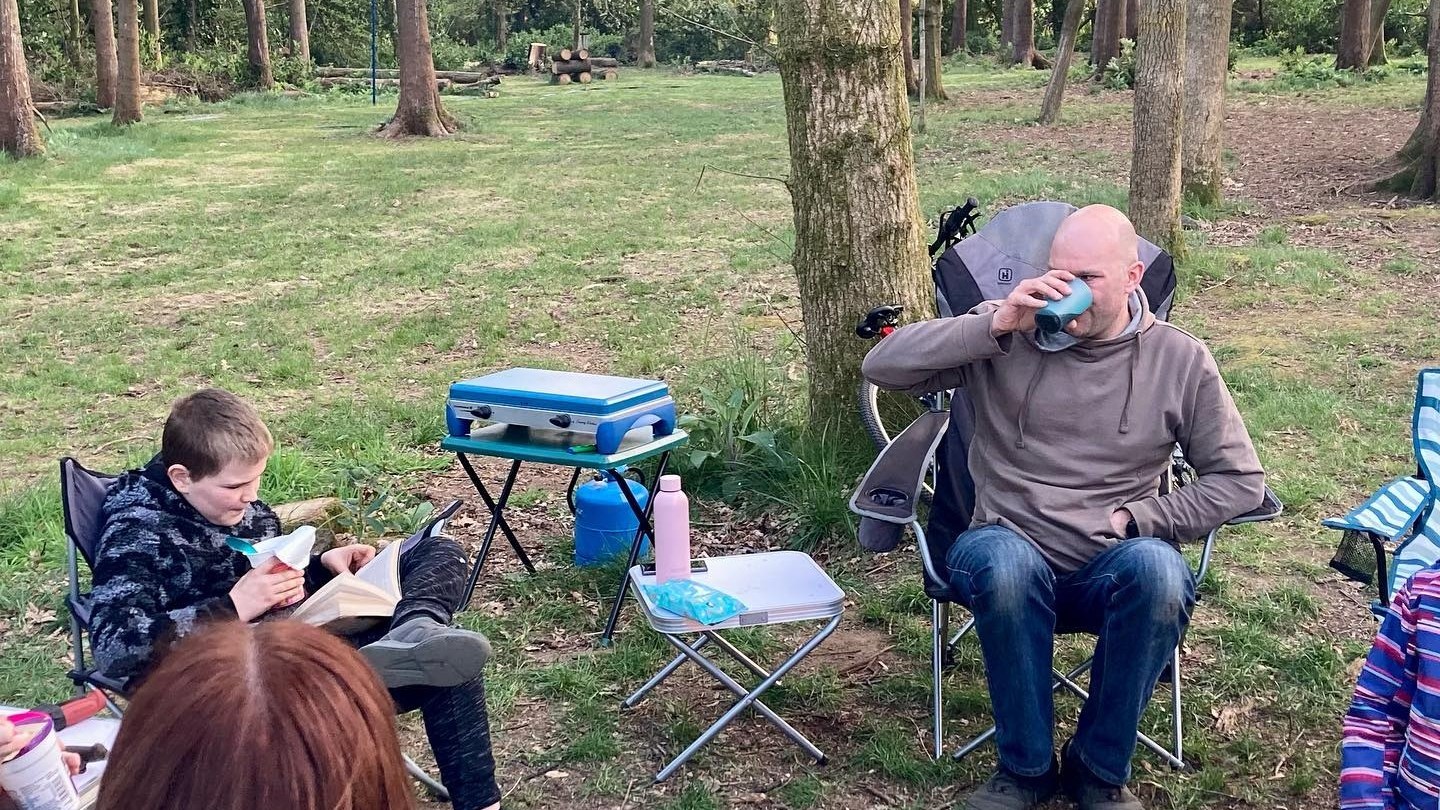People sitting around a campfire in a forest, one person reading a book, others relaxing in chairs.