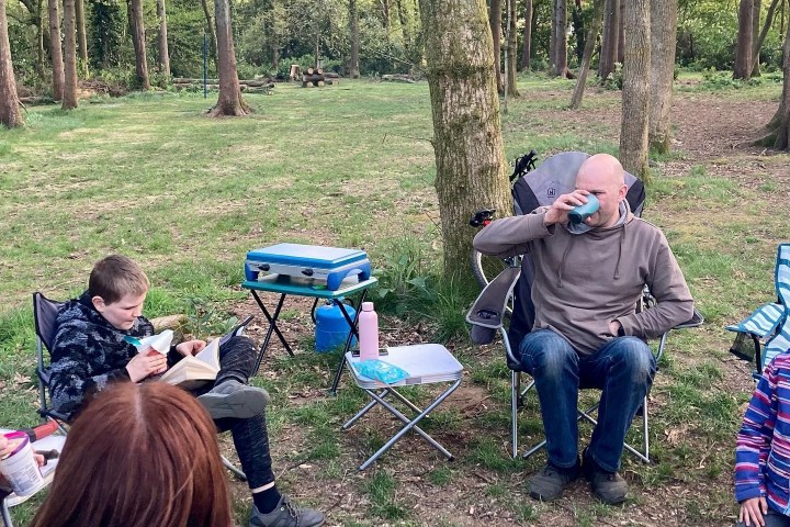 People sitting around a campfire in a forest, one person reading a book, others relaxing in chairs.