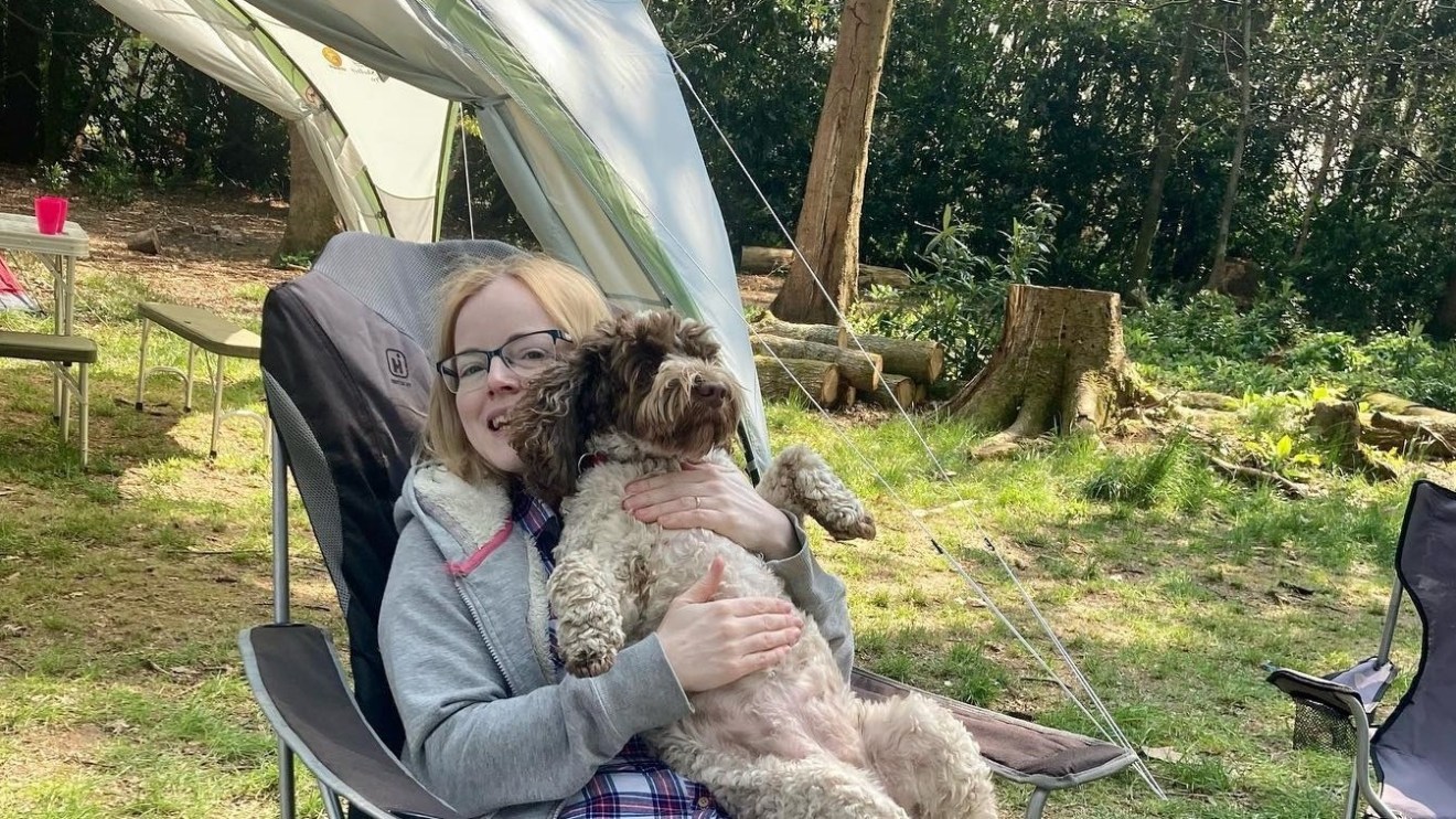 Person sitting in a camping chair holding a small dog in a wooded campsite.