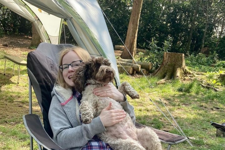 Person sitting in a camping chair holding a small dog in a wooded campsite.