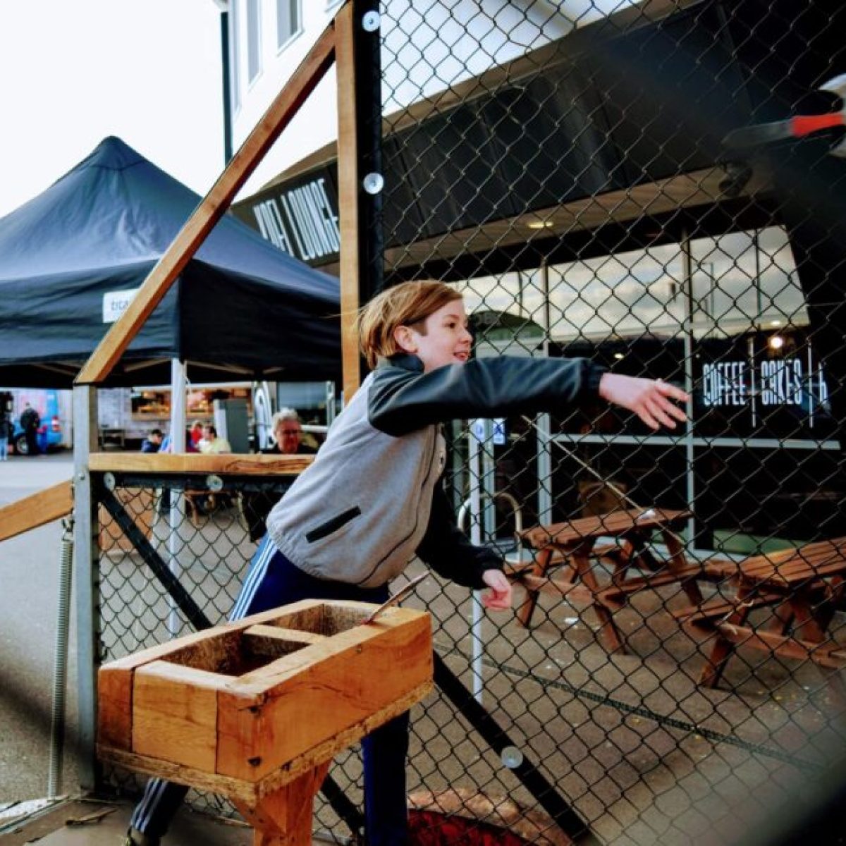 Child throwing an axe at an outdoor target near a chain-link fence and tables.