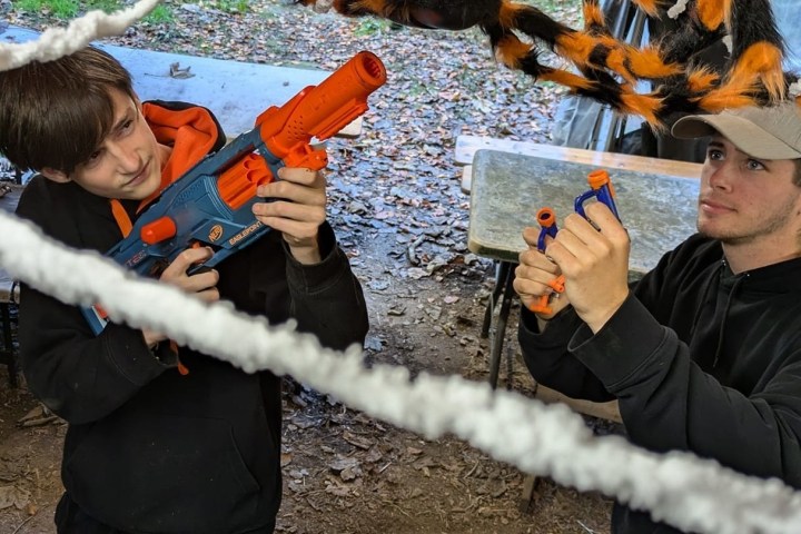 Two people aiming toy guns indoors with spider web decorations overhead.