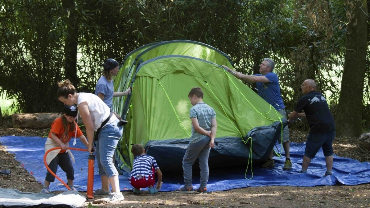 Group of people setting up a green tent in a wooded area on a blue tarp.