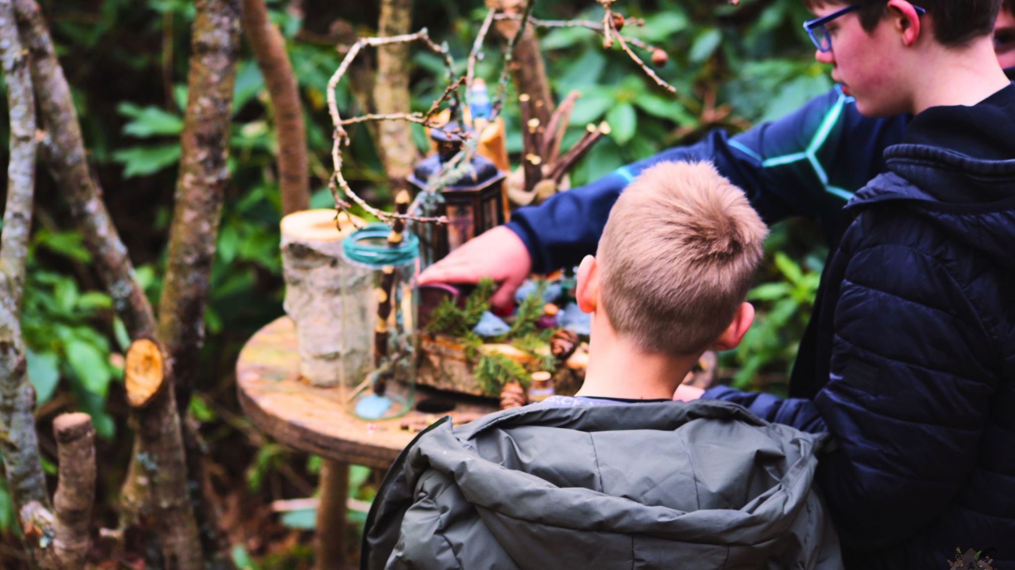 Two boys examining objects on a wooden table in a forest setting.