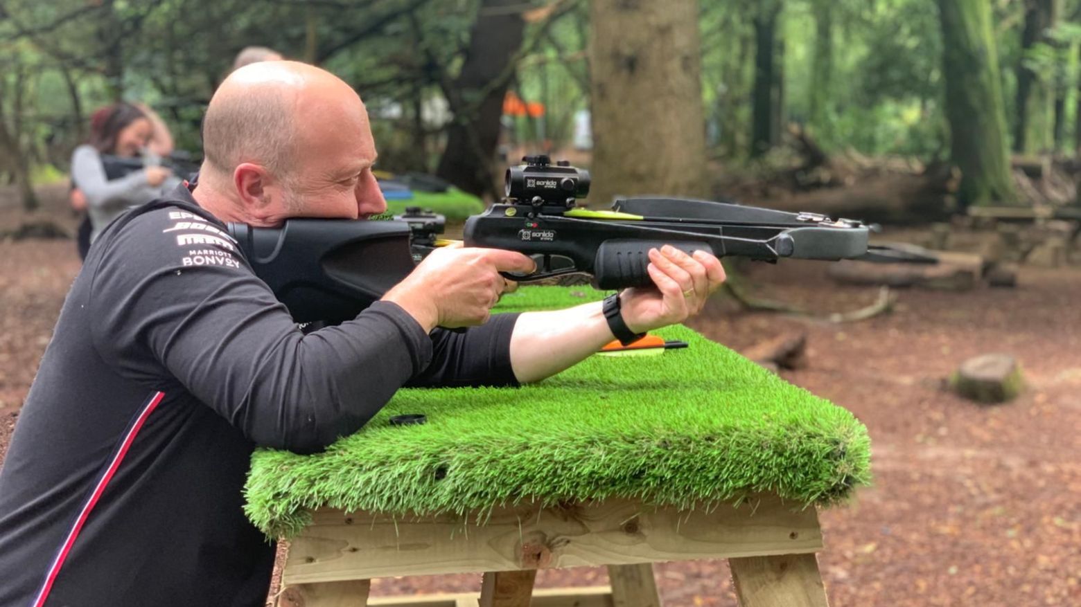 Man aiming a crossbow on a grassy stand in a wooded area.