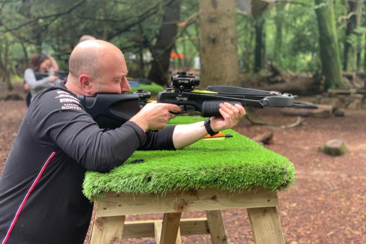 Man aiming a crossbow on a grassy stand in a wooded area.