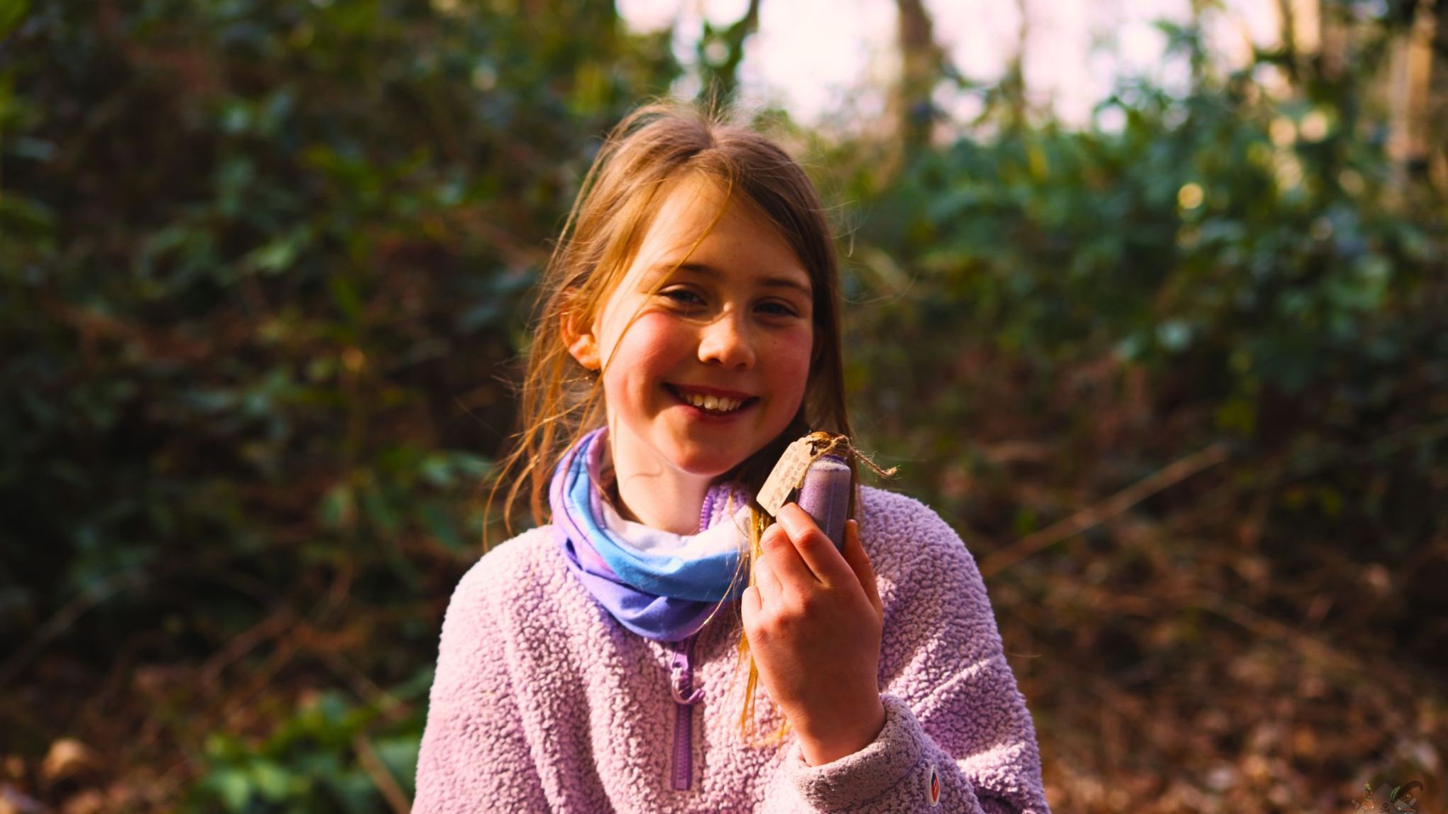 Smiling girl in a purple sweater holding a snack in a forest setting.
