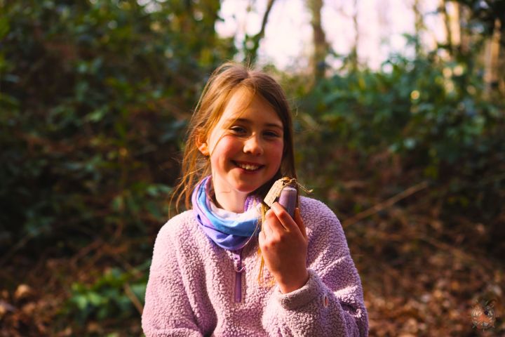 Smiling girl in a purple sweater holding a snack in a forest setting.