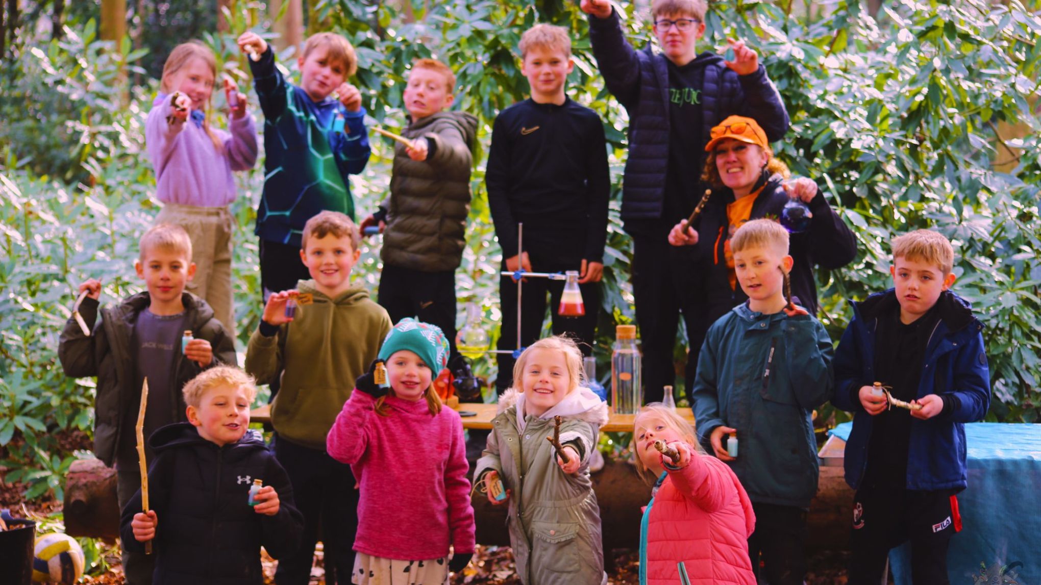Group of children and an adult holding sticks in a forest setting, smiling towards the camera.