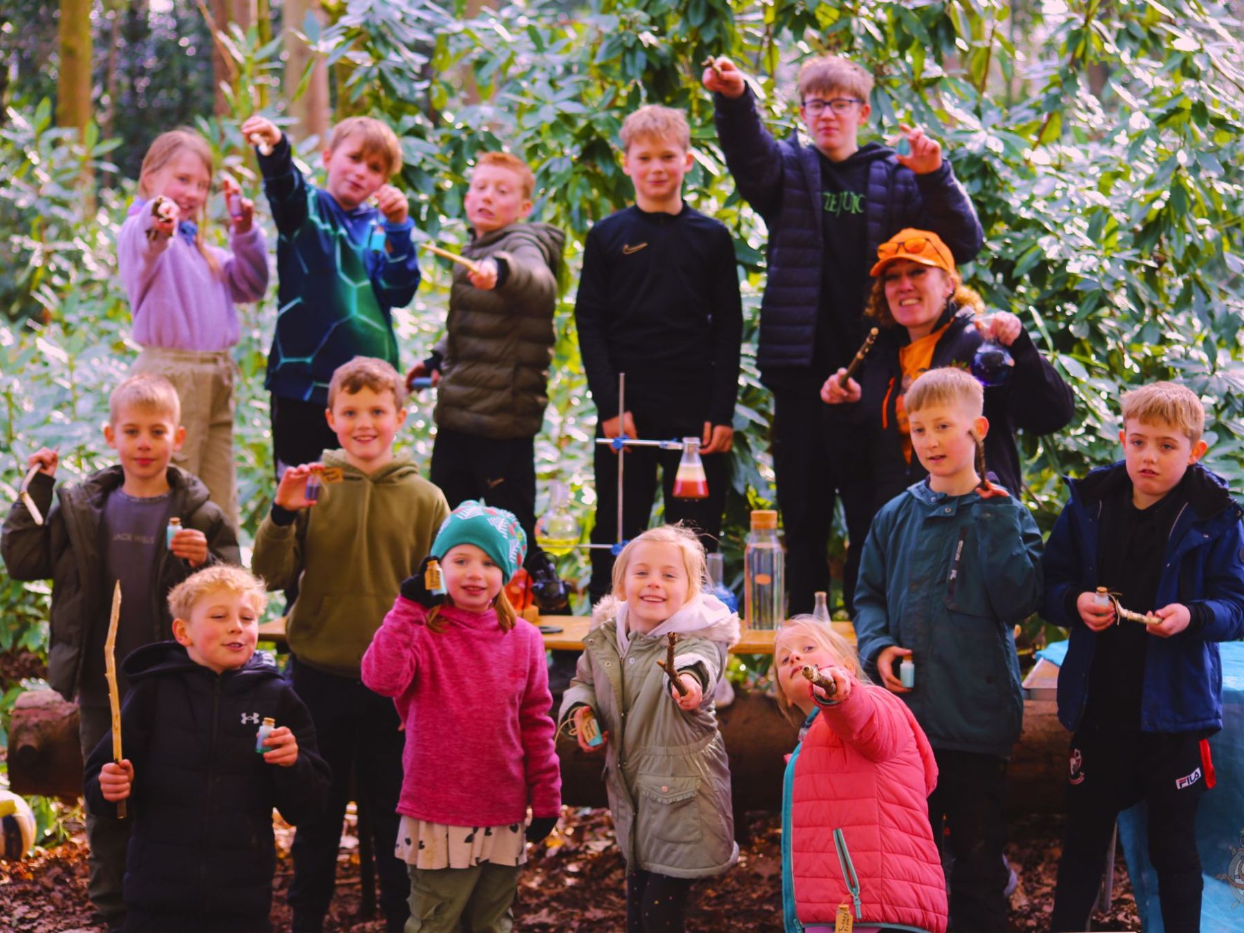 Group of children and an adult holding sticks in a forest setting, smiling towards the camera.