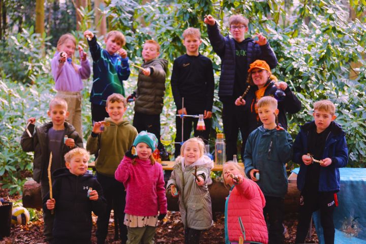 Group of children and an adult holding sticks in a forest setting, smiling towards the camera.