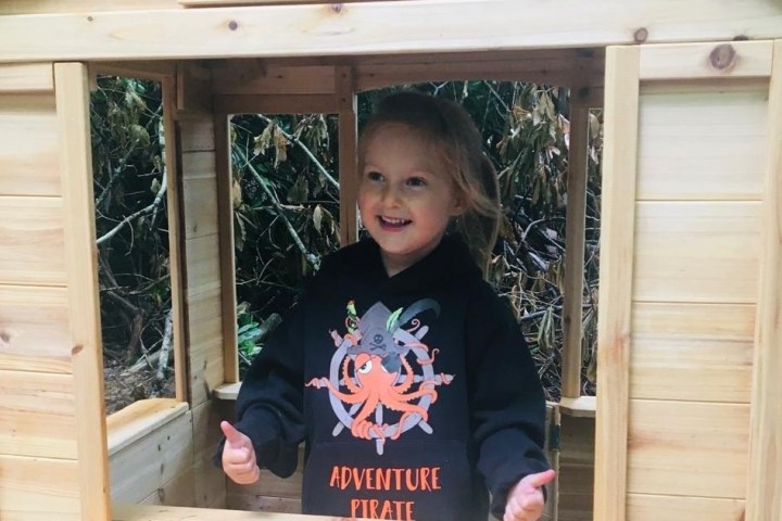 Child in black hoodie smiles while standing inside a wooden playhouse outdoors.