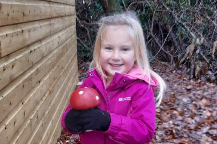 Child in purple coat holding a toy mushroom, standing next to a wooden wall in a forest.