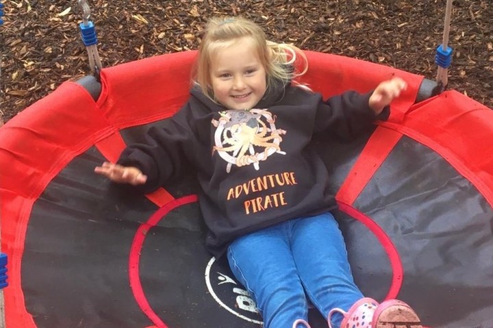 Child smiling and sitting on a round swing, wearing a hoodie with 'Adventure Pirate' text.